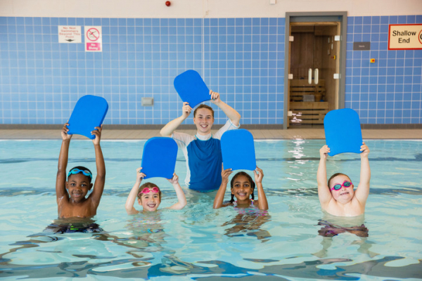 Four children in the shallow end of the pool having a swimming lesson with their instructor; children are standing in the water holding up blue kick boards